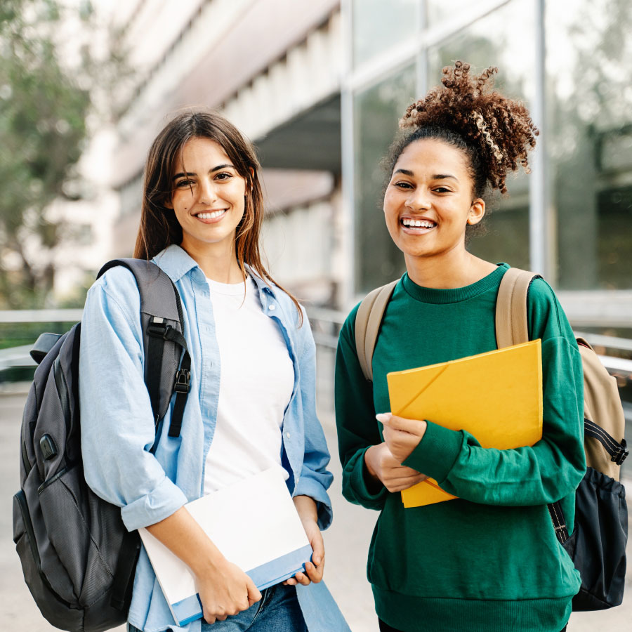 Two college students with backpacks smiling and holding books and folders